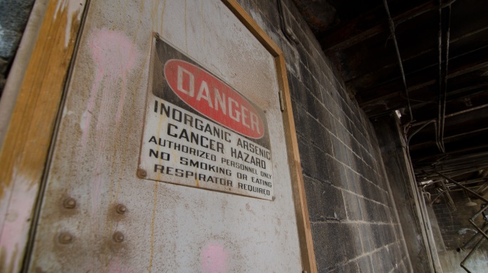 Dirty metal door with pink paint splatters and a sign that reads "Danger: Inorganic arsenic cancer hazard authorized personnel only no smoking or eating respirator required".